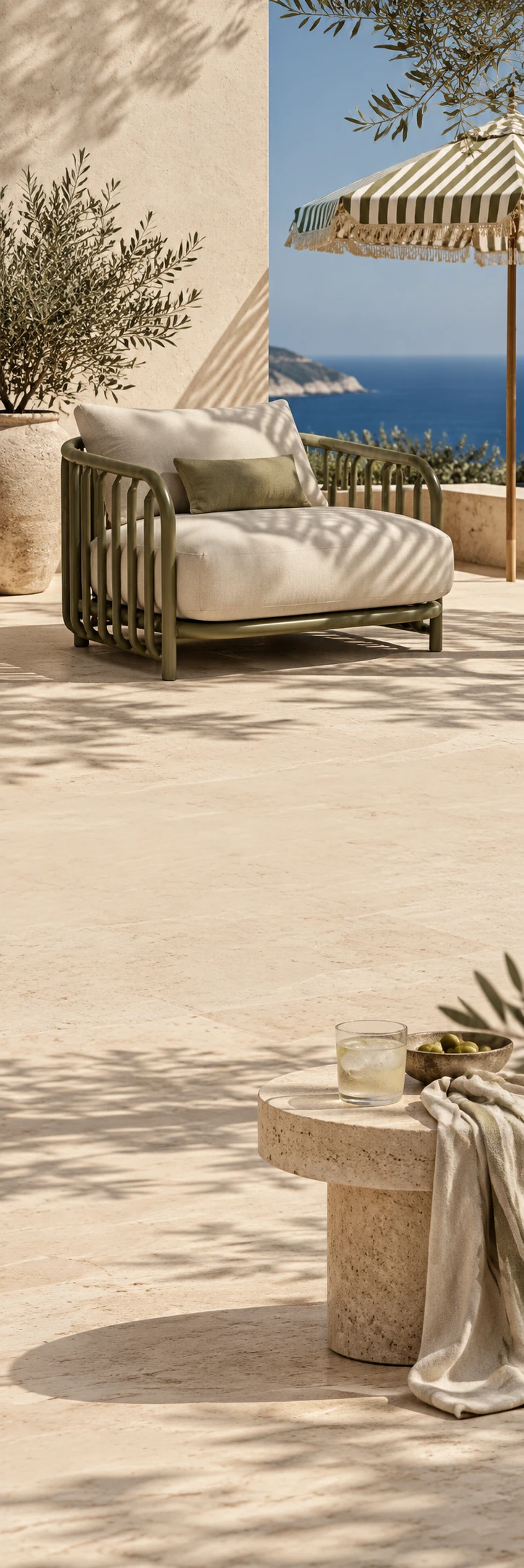 Quiet Mediterranean outdoor living editorial: sculptural olive-green garden lounge chair with oat linen cushion on pale travertine terrace, striped umbrella shadow, silver olive leaves at edge, deep blue sky softly visible. Lower scene warm stone side table with a cold glass and linen drape, same terrace continuing. Calm pale sandy copy spaces, hard sophisticated summer sunlight, no people.